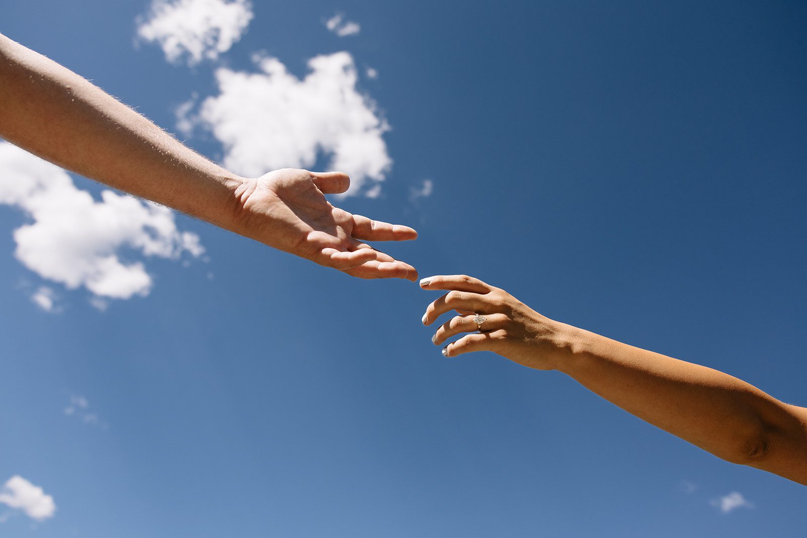 Two hands reach for eachother in the sky. One a male and one a female with an engagement ring on her ring finger